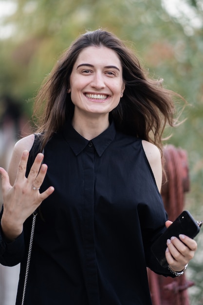 femme aux cheveux brune en vêtements sombres et lunettes de soleil. Photographie de rue de mode. Le mannequin est debout sur un pont suspendu et montre un anneau sur fond de nature. elle a dit oui