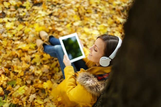 La femme aux cheveux bruns en manteau jaune et jeans assis et écoutant de la musique sous un arbre avec une tablette dans les mains et des écouteurs dans le parc de la ville d'automne par une chaude journée. Feuilles d'or d'automne. Vue de dessus