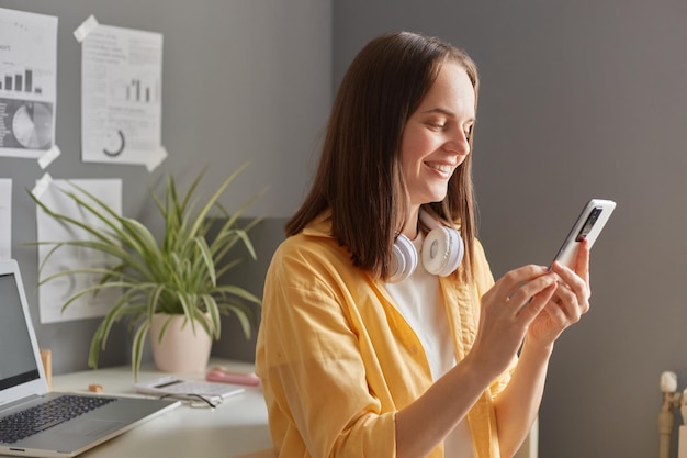 Une femme aux cheveux bruns prend une pause dans son bureau et sourit en regardant son téléphone portable se détendre avant de se remettre au travail en gérant son propre travail à distance