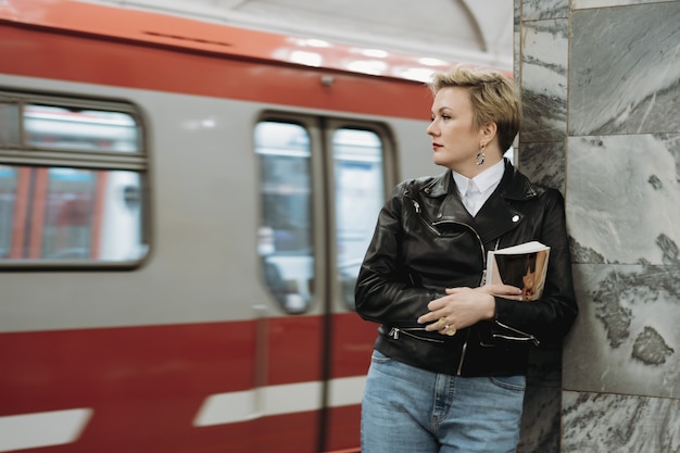 Femme aux cheveux courts en blouson de cuir avec livre en attente de train sur la plate-forme de la station de métro