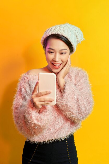 Femme aux cheveux courts excitée tenant un smartphone sur fond orange. Photo d'intérieur d'un merveilleux modèle féminin posant avec un téléphone.