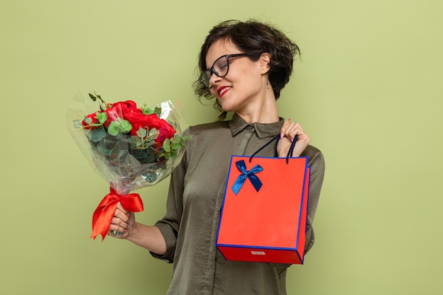 Photo femme aux cheveux courts tenant un bouquet de fleurs et un sac en papier avec des cadeaux souriant heureux et heureux célébrant la journée internationale de la femme le 8 mars debout sur fond vert