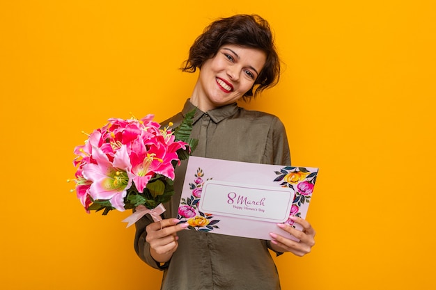 Femme aux cheveux courts tenant une carte de voeux et un bouquet de fleurs à l'air heureux et heureux souriant joyeusement célébrant la journée internationale de la femme le 8 mars