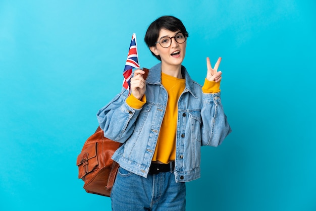 Femme aux cheveux courts tenant un drapeau du Royaume-Uni isolé sur mur bleu souriant et montrant le signe de la victoire