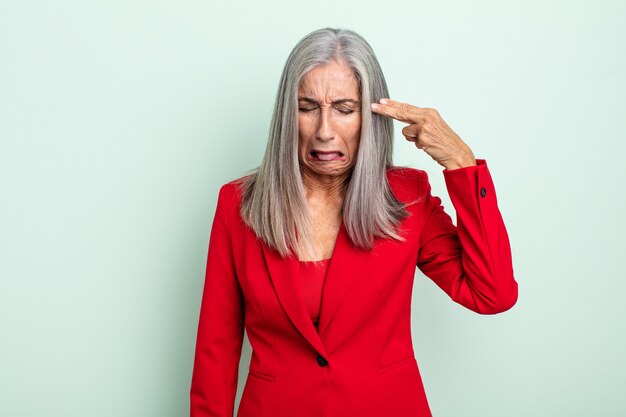 Femme aux cheveux gris d'âge moyen ayant l'air malheureuse et stressée, geste de suicide faisant signe d'arme à feu. concept de femme d'affaires senior