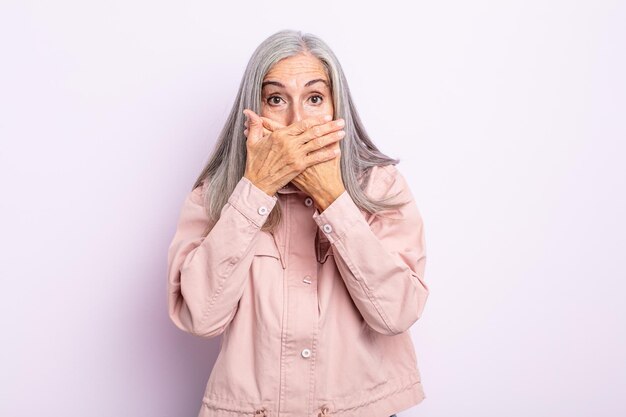 Femme aux cheveux gris d'âge moyen couvrant la bouche avec les mains avec une expression choquée et surprise, gardant un secret ou disant oups