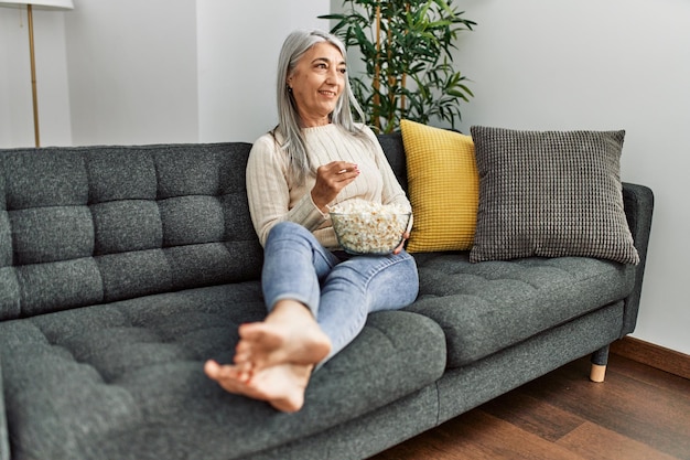Femme aux cheveux gris d'âge moyen souriant confiant regardant un film à la maison