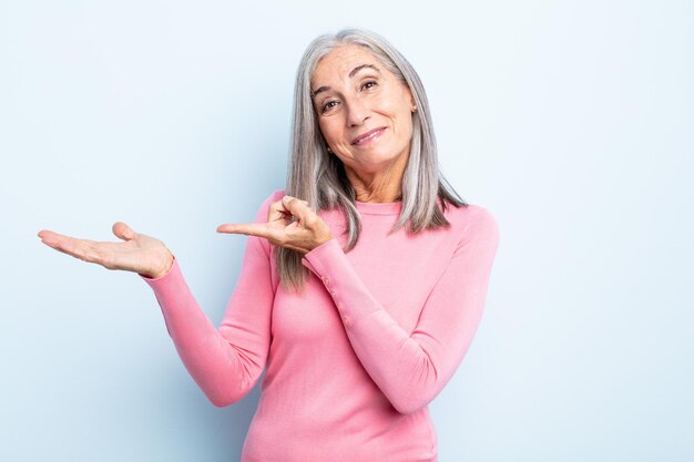 Femme aux cheveux gris d'âge moyen souriant joyeusement et pointant pour copier l'espace sur la paume sur le côté, montrant ou faisant la publicité d'un objet