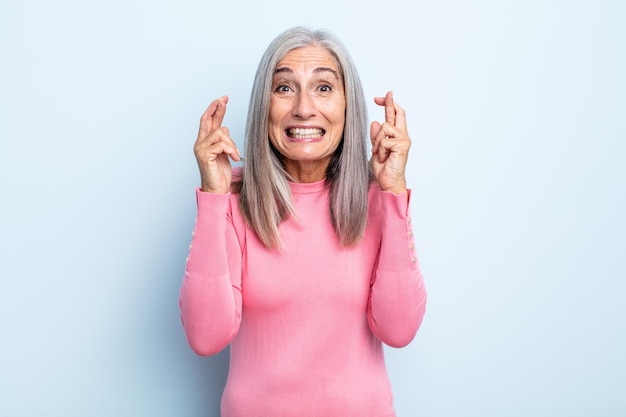 Femme aux cheveux gris d'âge moyen souriante et croisant anxieusement les deux doigts, se sentant inquiète et souhaitant ou espérant bonne chance