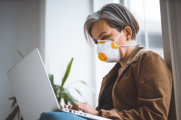 Femme aux cheveux gris pensif dans des vêtements décontractés et un masque de protection fonctionne sur un ordinateur portable assis sur le rebord de la fenêtre