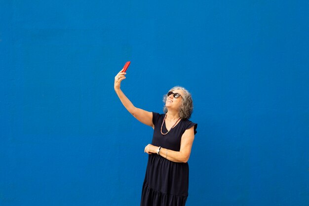 Femme aux cheveux gris senior prenant un selfie avec un smartphone rouge contre un mur bleu