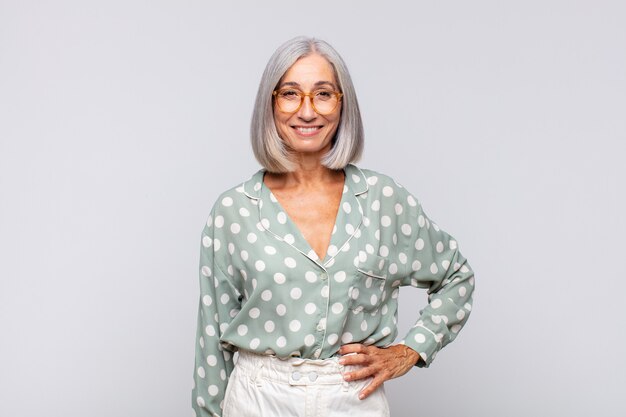 Femme aux cheveux gris souriant joyeusement avec une main sur la hanche et confiant isolé