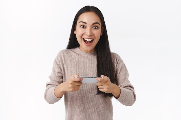 femme aux cheveux longs noirs, tenant le smartphone horizontalement, réussir, gagner le jeu, regarder l'appareil photo impressionné