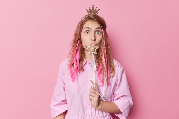 Une femme aux cheveux longs surprise couvre la bouche avec une baguette magique en forme d'étoile semble choquée par la caméra porte une couronne et la robe se sent impressionnée isolée sur fond rose Princesse prête à réaliser vos rêves