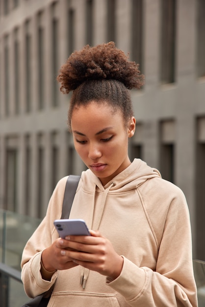 Photo une femme aux cheveux peignés tient une notification de contrôle de smartphone moderne lit un message sms vêtue d'un sweat-shirt beige porte un tapis de fitness se tient à l'extérieur