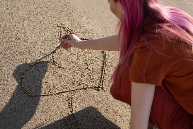 femme aux cheveux roses dessine un coeur avec un bâton sur le sable