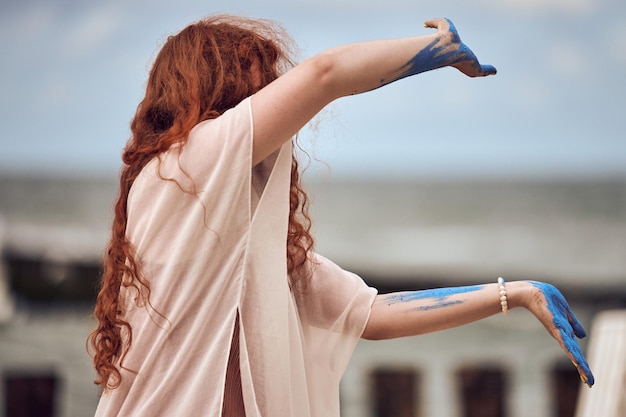 Femme aux cheveux rouges dansant sur la plage de la mer Performance artistique en plein air Fille rousse aux mains couvertes de peinture bleue dansant sur de la musique sur la côte de la mer Belle danse méditative sur le festival d'art