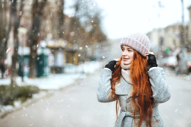 Une femme aux cheveux rouges émotive porte un manteau marchant dans la ville pendant les chutes de neige. Espace pour le texte