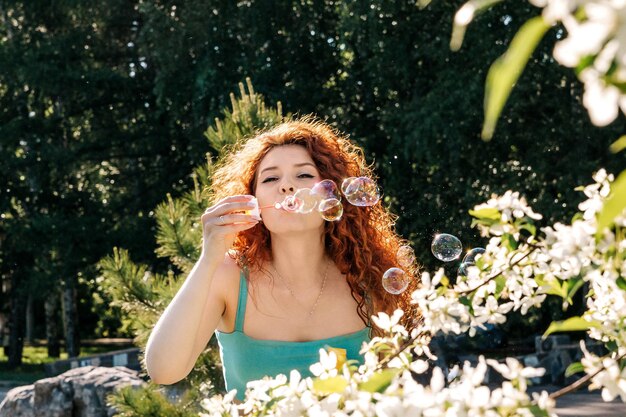 Une femme aux cheveux rouges et luxuriants souffle des bulles de savon au printemps pendant la floraison des pommiers