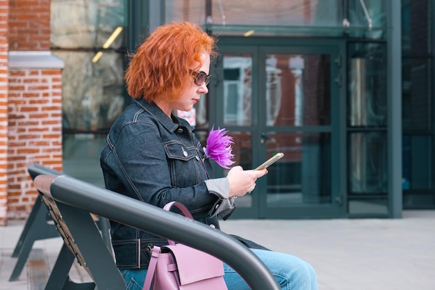 Une femme aux cheveux roux et aux lunettes de soleil tient un téléphone et regarde son téléphone.