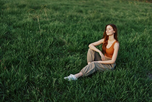 Photo la femme aux cheveux roux est assise dans le parc sur l'herbe verte portant un pantalon vert orange et des baskets et regarde le soleil d'été couchant photo de haute qualité