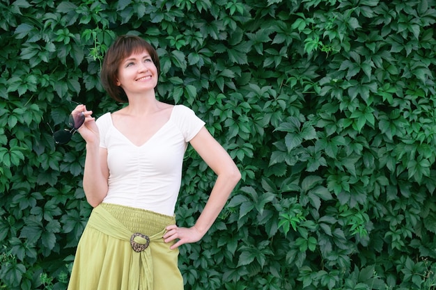 Femme aux cheveux roux foncé se dresse et sourit devant un mur couvert de feuilles