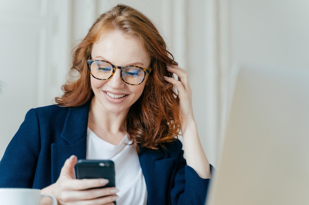 Une femme aux cheveux roux satisfaite utilise un appareil électronique moderne pour vérifier la communication en ligne du fil d'actualité et rechercher des informations se trouve dans un espace de coworking utilise une connexion Internet haut débit