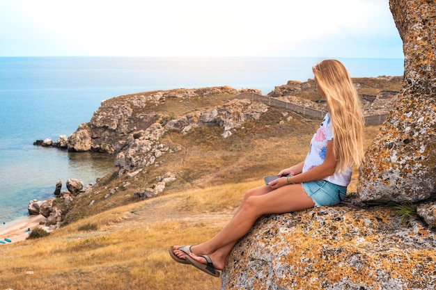 Une femme aux longs cheveux blonds est assise sur un rocher sur une montagne surplombant un littoral rocheux Voyage et tourisme