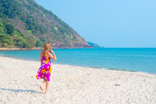 Une femme aux longs cheveux blonds vêtue d'une robe brillante se promène le long de la plage de sable sur une île tropicale, vue arrière.