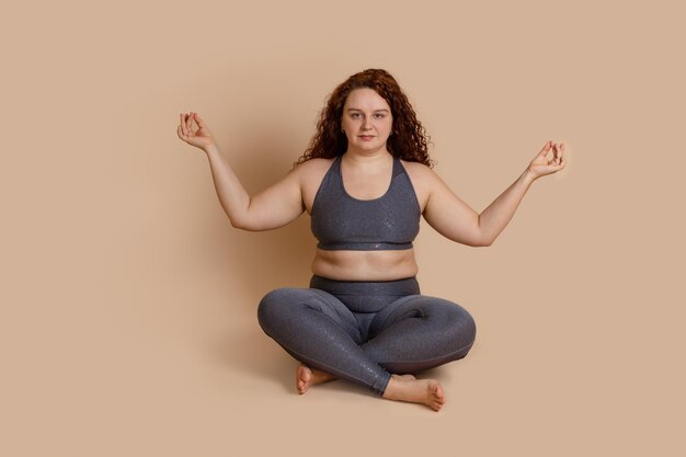 Femme aux pieds nus calme et joyeuse aux cheveux bouclés assise dans un lotus ashtanga asana position de yoga croisant les jambes en méditant portant des vêtements de sport gris haut et sportif Problèmes avec le traitement de rotation des os