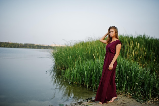 Femme aux pieds nus sensuelle blonde en robe de marsala rouge posant contre le lac avec des roseaux