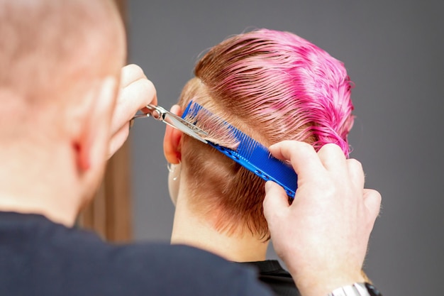 Femme ayant une nouvelle coupe de cheveux. Coiffeur masculin coupant les cheveux courts roses avec des ciseaux dans un salon de coiffure.