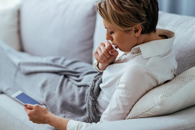 Femme ayant le virus du rhume et de la grippe et toussant en utilisant un téléphone portable sur le canapé