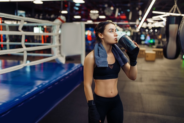 Femme en bandages de boxe noirs boit de l'eau sur le ring, formation de boîte. Boxer dans la salle de sport, kickboxer fille dans un club de sport, séance d'entraînement de kickboxing