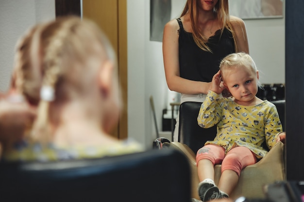 Une femme de barbier fait une jolie coiffure à la mode pour une jolie petite fille blonde dans un salon de coiffure moderne, un salon de coiffure. Le coiffeur fait la coiffure pour le jeune bébé dans le salon de coiffure. Concept coiffure et beauté