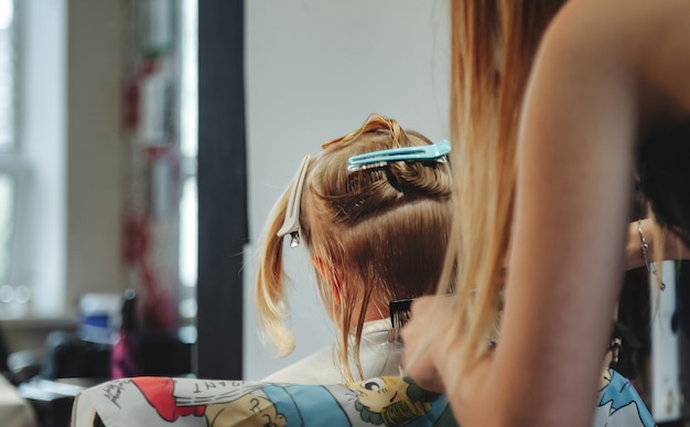 Une femme de barbier fait une jolie coiffure à la mode pour une jolie petite fille blonde dans un salon de coiffure moderne, un salon de coiffure. Le coiffeur fait la coiffure pour le jeune bébé dans le salon de coiffure. Concept coiffure et beauté