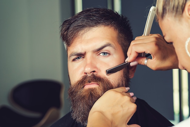 Femme de barbier rasant la barbe du client dans un salon de coiffure ou un salon de beauté. Portrait d'un client masculin se rasant la barbe au salon. Barbier au travail. Homme au salon de coiffure. Bel homme avec barbe en salon de coiffure.