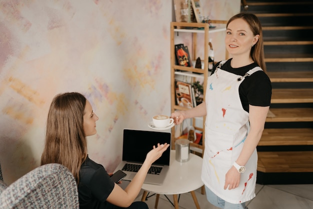 Une femme barista avec piercing face tend un café au lait à une fille dans un café. Une jeune femme aux cheveux longs travaillant à distance sur un ordinateur portable maintient la distance sociale attrape une tasse de café dans un café.