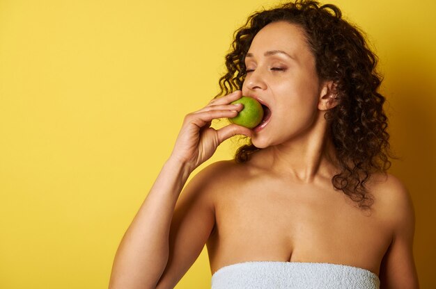 Une femme basanée aux cheveux bouclés mord une pomme verte en se tenant debout sur un fond jaune.