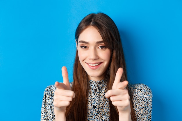 Femme de beauté excitée et confiante, pointant du doigt la caméra et souriant, vous invite et vous fait signe, rejoignez-nous geste, louant le bon choix, debout sur le bleu.