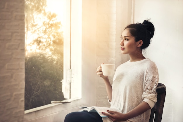 femme de beauté avec livre et tasse de café assis regardant par la fenêtre