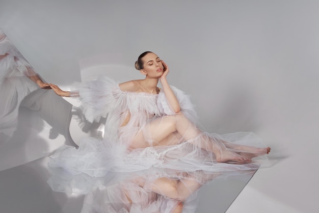 Une femme de beauté romantique vêtue d'une robe transparente légère se reflète dans un miroir flexible assis sur le sol. Corps parfait et longues jambes