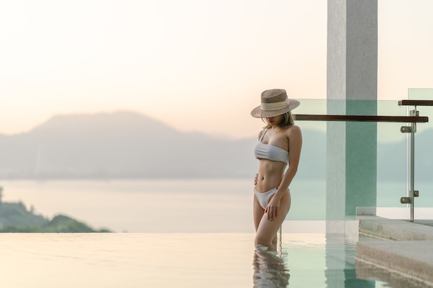 Femme en bikini blanc marchant pose près de la piscine avec le balcon transparent en verre et vue sur l'océan.