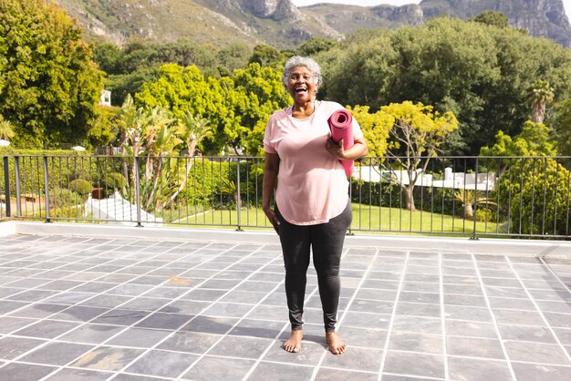 Une femme biraciale âgée aux cheveux gris tient un tapis de yoga rose et sourit à l'extérieur.