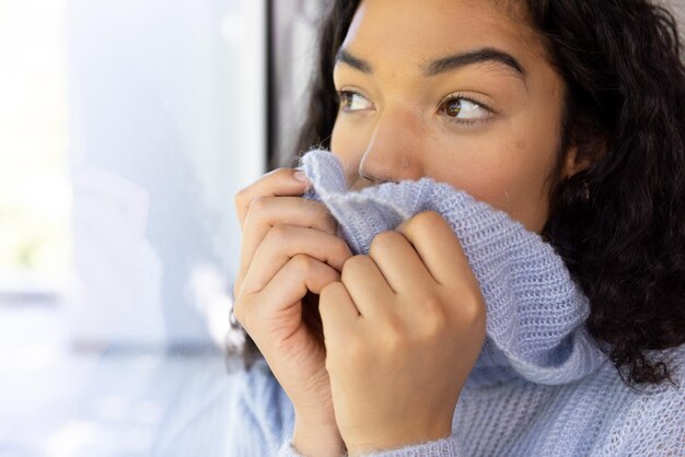 Photo une femme biraciale se couvrant d'un pull et regardant par la fenêtre vers une maison ensoleillée.