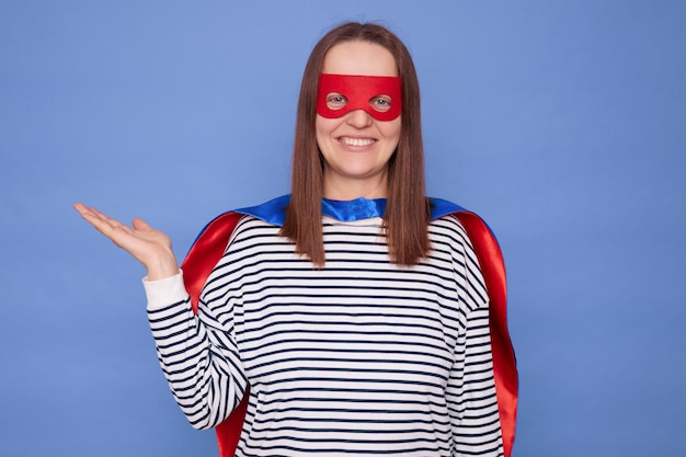 Une femme blanche aux cheveux bruns souriante et ravie portant un costume de super-héros et une chemise à rayures étendant la paume avec un espace de copie pour le texte promotionnel de la publicité isolé sur un fond bleu.