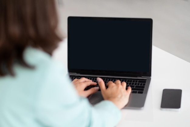 Une femme blanche du millénaire tapant sur un ordinateur avec un écran vide sur le lieu de travail avec un smartphone