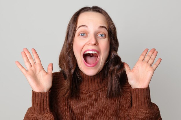 Une femme blanche extrêmement heureuse portant un pull brun se tient isolée sur un fond gris clair en levant les bras en criant d'émotions positives en se réjouissant