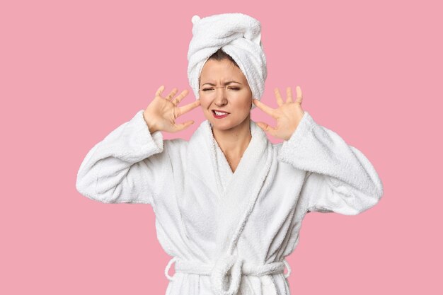 Une femme blanche en peignoir et serviette couvrant les oreilles avec des doigts stressés et désespérés