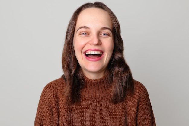 Une femme blanche riante portant un pull brun se tient isolée sur un fond gris clair est de bonne humeur entend une blague drôle exprimant des émotions positives